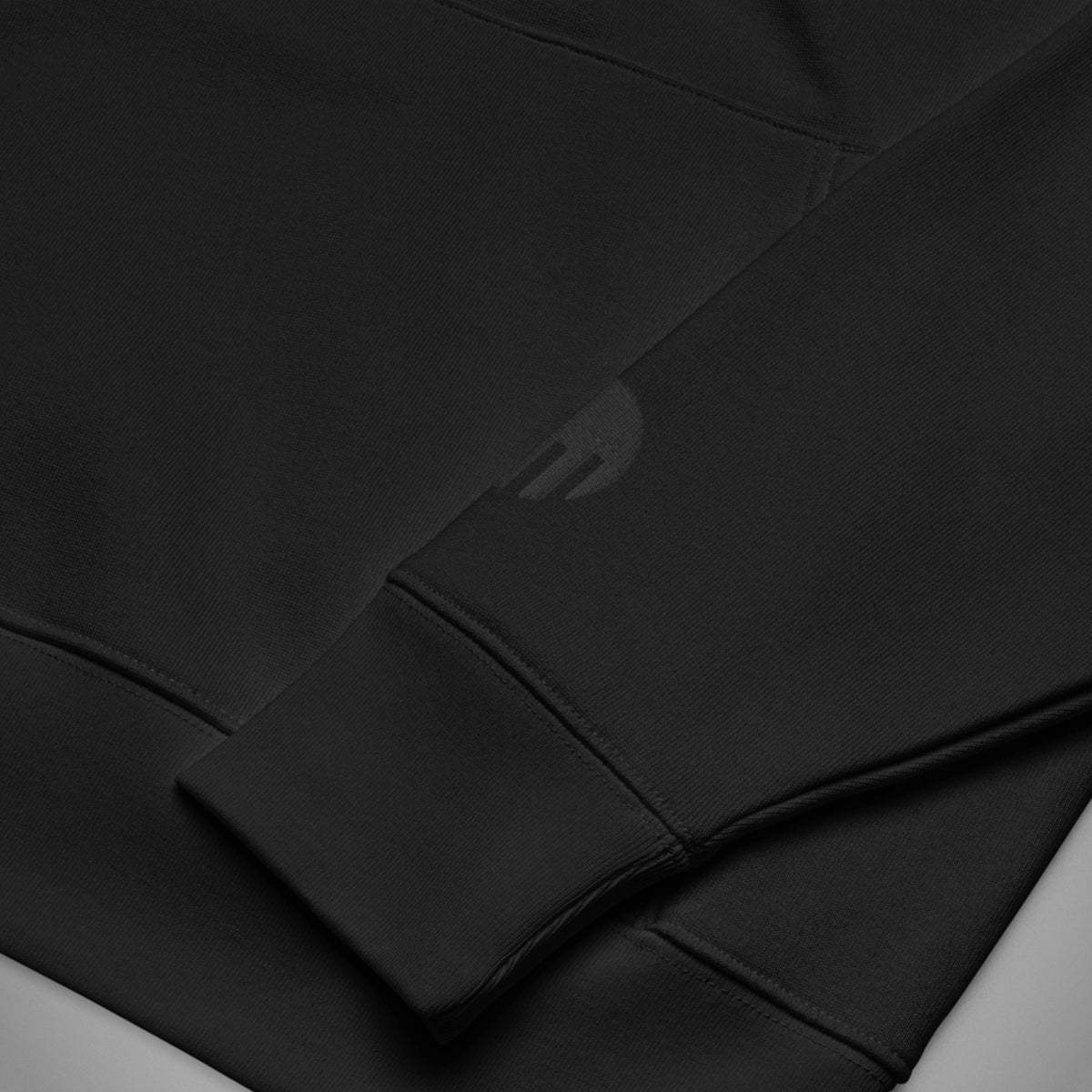 Eine Nahaufnahme des linken Ärmels eines schwarzen Regular Fit Hoodies der Marke FORCE ZERO im Black Stealth Design zeigt das präzise, rund gestickte FORCE ZERO Logo dezent Ton-in-Ton in Schwarz oberhalb des gerippten Ärmelbündchens.