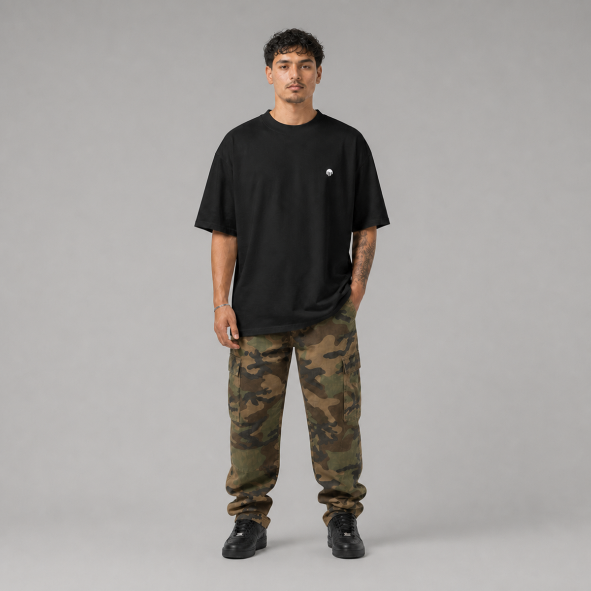 Ein schwarzes Oversized T-Shirt der Marke FORCE ZERO im Rogue Mode Raw Design am Model in der Produkt-Vorderansicht in der Totalen mit einem minimalistischen, kleinen weißen Logo-Print auf der linken Brustseite, kombiniert mit einer Camouflage-Cargo-Hose.