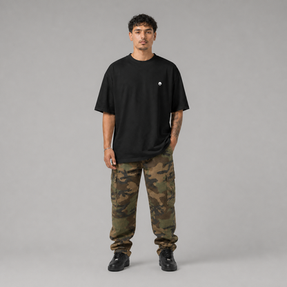 Ein schwarzes Oversized T-Shirt der Marke FORCE ZERO im Rogue Mode Raw Design am Model in der Produkt-Vorderansicht in der Totalen mit einem minimalistischen, kleinen weißen Logo-Print auf der linken Brustseite, kombiniert mit einer Camouflage-Cargo-Hose.