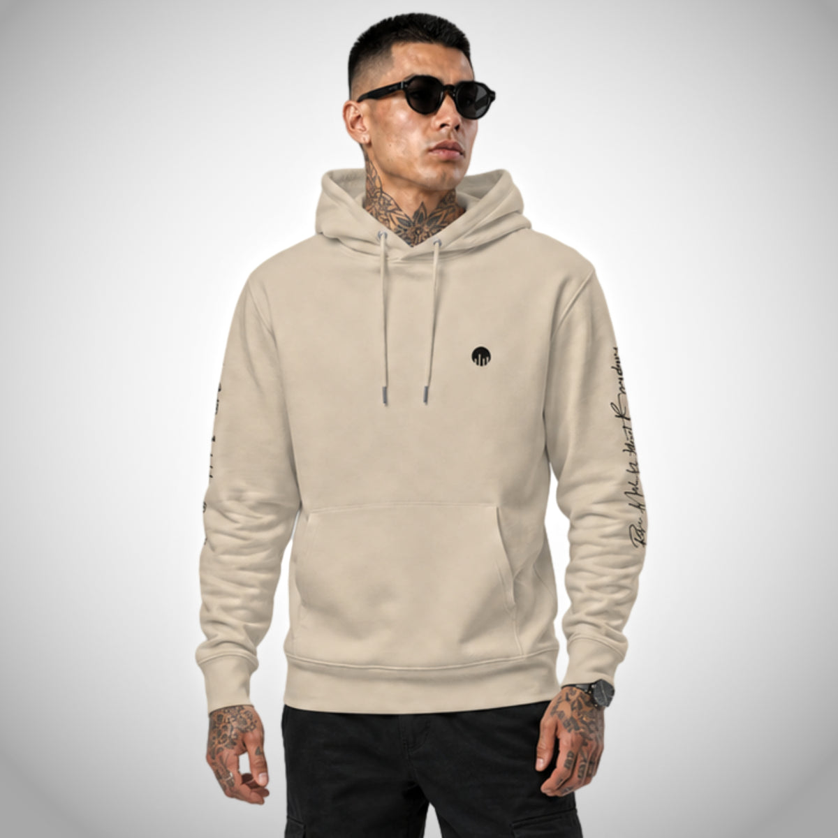 Ein beiger Core Fit Hoodie der Marke FORCE ZERO im Rogue Mode Renegade Design zeigt als Ganzkörper-Modellbild die Vorderansicht an einem tätowierten Model mit Sonnenbrille und schwarzer Hose, wobei auf der linken Brust das runde schwarze Logo-Emblem und auf dem linken Ärmel ein schwarzer Schriftzug-Print zu sehen sind.