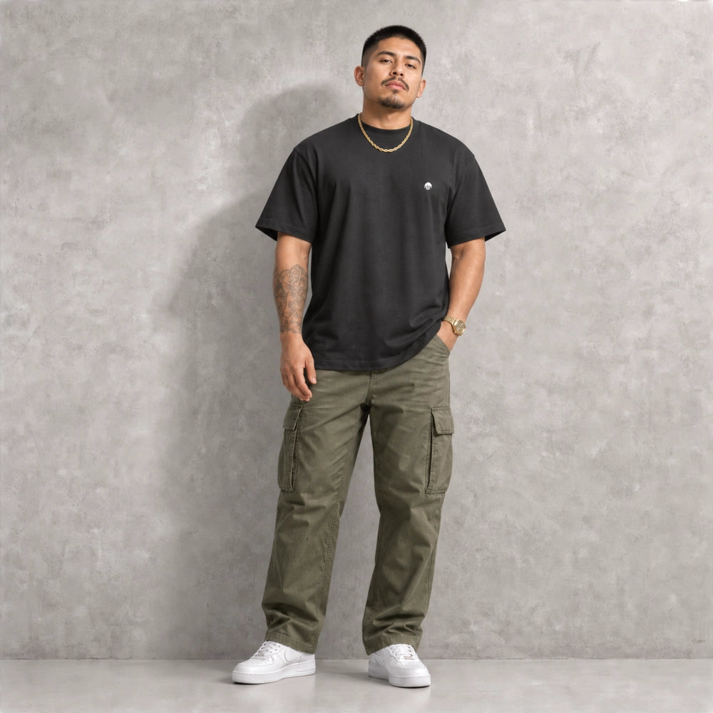 Ein schwarzes Boxy Cut T-Shirt der Marke FORCE ZERO im Rogue Mode Renegade Design zeigt als Ganzkörper-Modellbild ein Model in Frontalansicht mit einer goldenen Halskette, grüner Cargo-Hose und weißen Sneakern vor einer grauen Betonwand, wobei auf der linken Brust der kleine, runde, weiße Logo-Print zu sehen ist.