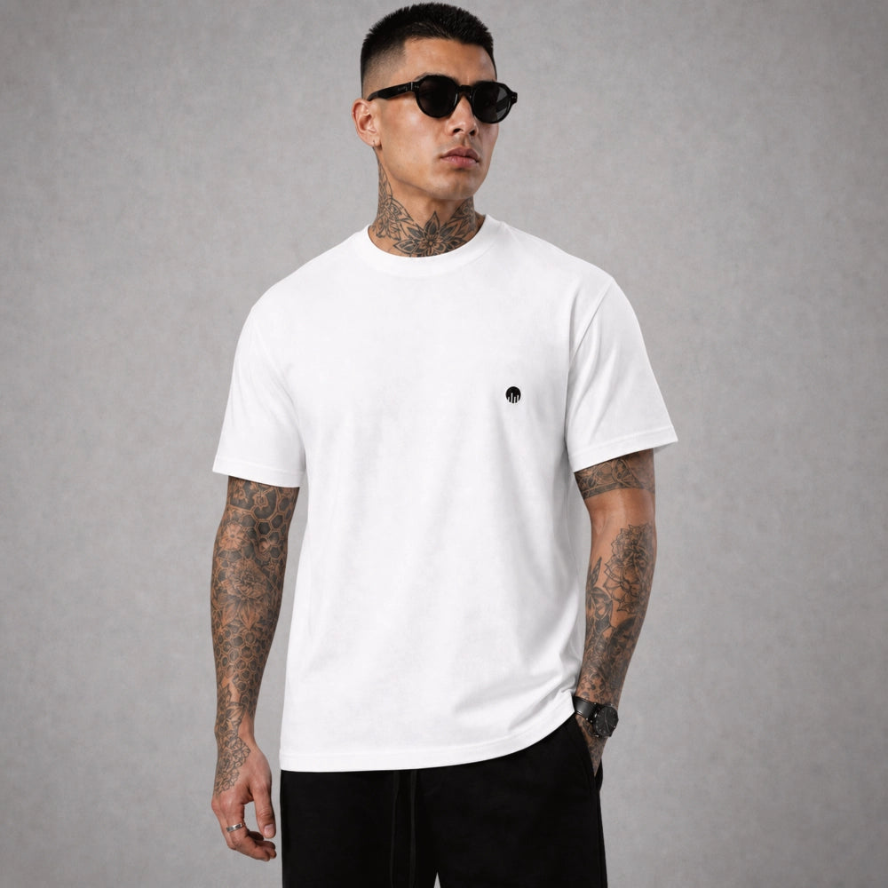 Ein weißes Boxy Cut T-Shirt der Marke FORCE ZERO im Rogue Mode Renegade Design zeigt als Ganzkörper-Modellbild eine leicht angewinkelte Seitenansicht an einem tätowierten Model mit Sonnenbrille und Blick zur Seite, wobei der kleine, runde, schwarze Logo-Print auf der linken Brust gut sichtbar ist.