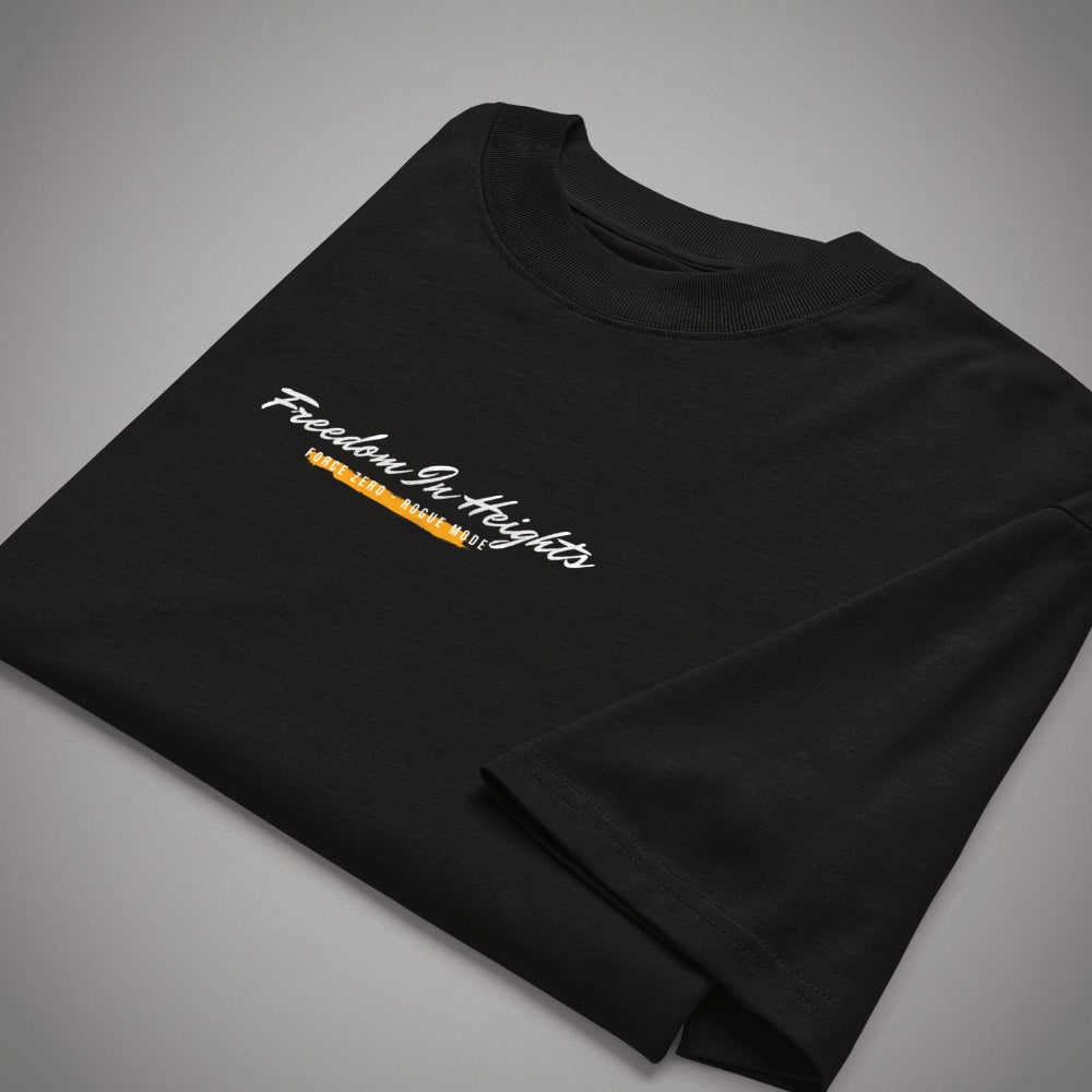 Ein gefaltetes schwarzes Oversized T-Shirt der Marke FORCE ZERO im Rogue Mode Rio X Design in der Produkt-Vorderansicht (Mockup) mit Fokus auf den geschwungenen weißen Brustprint „Freedom In Heights“ über einer gelb-orangefarbenen, markierten Grafikelement-Unterzeilung.