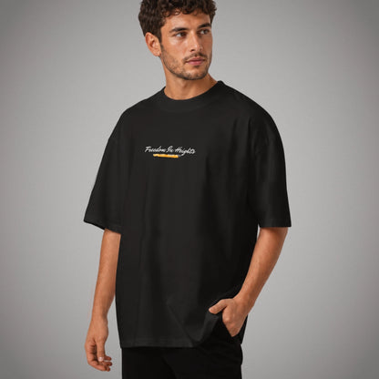 Ein schwarzes Oversized T-Shirt der Marke FORCE ZERO im Rogue Mode Rio X Design am Model in der Produkt-Vorderansicht mit dem geschwungenen weißen Brustprint „Freedom In Heights“ über einer gelb-orangefarbenen, markierten Grafikelement-Unterzeilung.